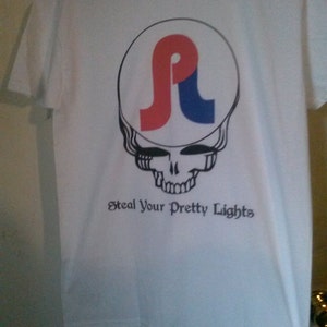 May include: White t-shirt with a black and white skull graphic and a red, white, and blue logo inside the skull. The text "Steal Your Pretty Lights" is printed below the skull.