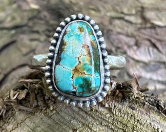 May include: A silver ring featuring a large, teardrop-shaped turquoise stone with prominent brown matrix veining. The stone is set in a serrated silver bezel, surrounded by a decorative beaded silver border, and attached to a textured silver band.