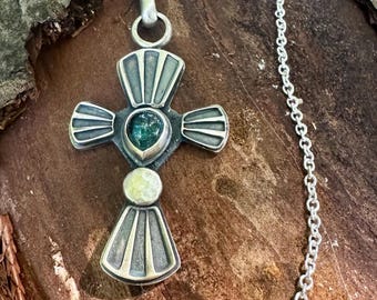 May include: A silver cross pendant necklace featuring a central teardrop-shaped green stone and a circular pale yellow stone. The cross has a textured, oxidized finish with radiating line patterns on each arm and hangs from a delicate silver chain.