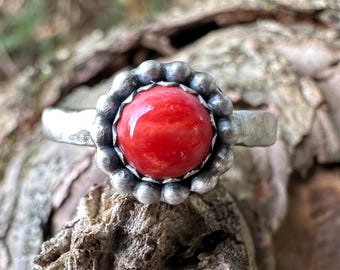 May include: A ring featuring a smooth, round, vibrant red stone set in a scalloped, oxidized silver bezel. The band of the ring has a hammered, textured finish, providing a contrast to the polished surface of the central red gemstone.