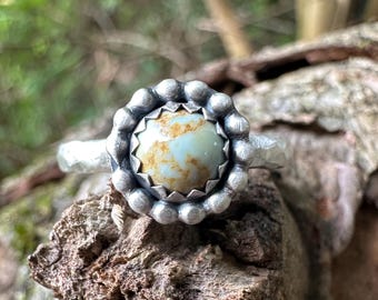 May include: A silver ring featuring a round, light blue and brown mottled stone set in a serrated bezel. The stone is surrounded by a decorative border of small, rounded silver spheres. The ring band has a textured, hammered finish.
