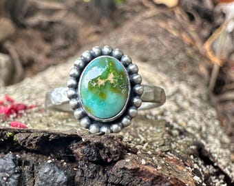 May include: A sterling silver ring featuring an oval-shaped turquoise stone with green and brown matrix patterns. The stone is set in a bezel surrounded by a decorative beaded border, mounted on a hammered silver band with a textured finish.