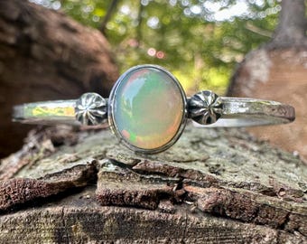 May include: A silver cuff bracelet featuring a central oval-shaped opal gemstone with iridescent flashes of color. The metal band has a hammered texture and is accented by two small, circular, star-shaped silver ornaments flanking the opal.