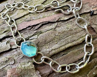 May include: A silver chain bracelet featuring a single hexagonal, faceted, translucent light blue stone set in a silver bezel. The bracelet is constructed with oval-shaped links and secures with a simple hook-and-eye clasp mechanism.