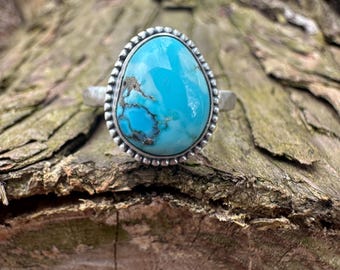 May include: A silver ring featuring a large, oval turquoise stone with brown veining. The stone is set in a beaded bezel. The ring is displayed on a textured, brown wooden surface.