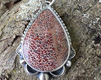 May include: A teardrop-shaped pendant featuring a reddish-brown and cream-colored patterned stone set in a silver frame. The metal setting includes a beaded border, decorative fan-shaped accents, and smooth silver spheres at the base.