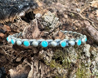 May include: A silver-toned bracelet with turquoise-colored stones. The bracelet has a twisted design and is set against a natural, textured background of wood and moss. The turquoise stones are evenly spaced around the bracelet.