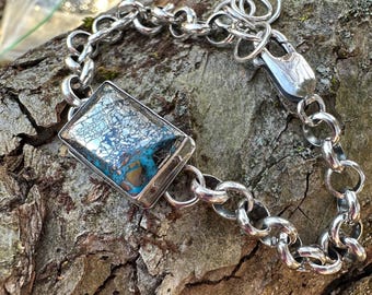 May include: A silver bracelet with a rectangular, patterned stone centerpiece. The stone features shades of blue, brown, and white. The bracelet has a chunky chain design and a secure clasp. The jewelry is displayed on a textured wooden surface.