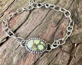 May include: A silver chain bracelet featuring a central oval-shaped stone with a light green and brown mottled pattern. The stone is set in a beaded silver bezel, and the bracelet is secured with a simple hook clasp at the end of the chain links.
