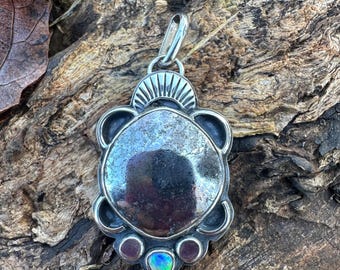 May include: A silver pendant with a large, reflective, oval-shaped center stone. The pendant has a decorative border and three smaller stones at the bottom, one of which has an iridescent sheen. The pendant is suspended from a silver bail.