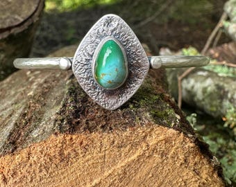 May include: A silver cuff bracelet with a textured, diamond-shaped centerpiece. The centerpiece holds a vibrant, oval-shaped turquoise stone. The bracelet is displayed on a wooden surface, showcasing its handcrafted details and natural aesthetic.