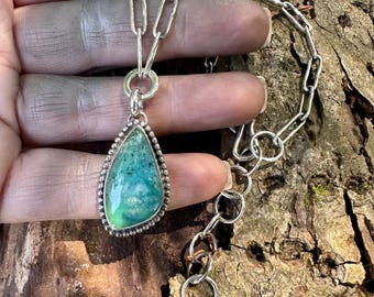 May include: A silver necklace featuring a teardrop-shaped green stone pendant with a beaded border. The pendant hangs from a textured silver chain with elongated oval links and a circular clasp, showcasing a polished finish on the stone surface.