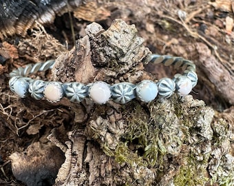 May include: A silver bracelet with a twisted band and a row of light pink, round stones. The stones are set between decorative silver elements. The bracelet is displayed on a textured, mossy surface.