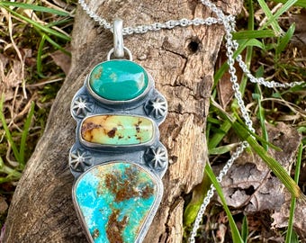 May include: A silver pendant necklace featuring three turquoise stones in varying shades of blue and green. The pendant has a unique design with star-shaped accents and is suspended from a delicate silver chain. The necklace is displayed on a piece of weathered wood.