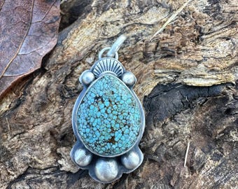 May include: A silver pendant with a large, oval turquoise stone. The stone is a vibrant blue-green color with a speckled pattern. The pendant has a decorative silver setting with small silver beads and a loop for a chain.