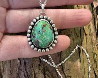 May include: A silver necklace featuring a pendant with a vibrant green stone. The stone is oval-shaped and surrounded by a beaded silver frame. The necklace chain is delicate and silver, creating a simple yet elegant design. The pendant's green color is eye-catching.