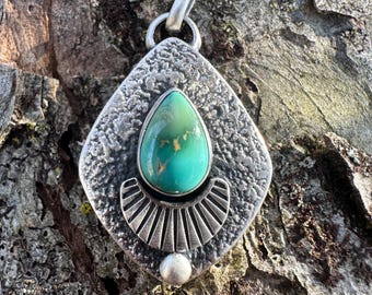 May include: A silver pendant with a turquoise gemstone. The pendant has a textured, diamond-shaped frame with a teardrop-shaped turquoise stone in the center. A fan-shaped design sits below the stone, and a small silver ball is at the bottom.