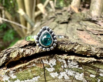 May include: A silver ring featuring a teardrop-shaped teal gemstone set in a bezel. The stone is surrounded by a decorative border of small silver spheres. The ring band has a hammered, textured finish with a polished, reflective surface.