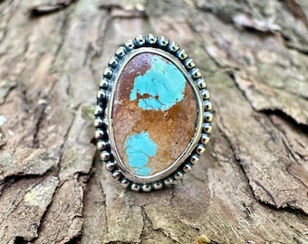 May include: A silver ring featuring an oval-shaped stone with a brown matrix and two distinct patches of bright turquoise blue. The stone is set in a polished silver bezel surrounded by a decorative border of small, rounded silver beads.