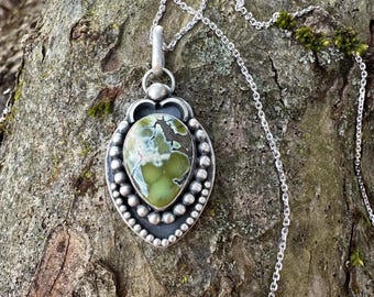 May include: A silver pendant necklace featuring a teardrop-shaped, green stone with a unique pattern. The stone is set in a detailed silver frame with a beaded border. The necklace chain is also silver.