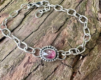 May include: A silver chain bracelet featuring a central oval-shaped gemstone with a faceted surface. The stone displays a gradient of pink and dark purple hues. The setting is decorated with a beaded border, and the bracelet includes a hook clasp.