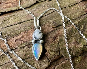 May include: A silver necklace featuring a pendant with two stacked stones. The top stone is a round, speckled white and gray gem, while the bottom stone is a teardrop-shaped, iridescent gem with blue and yellow hues, set in a silver frame.