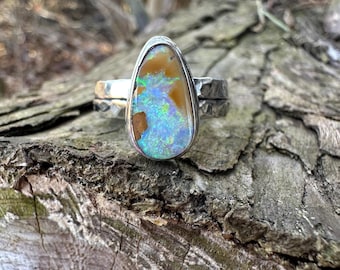 May include: A silver ring featuring a large, teardrop-shaped opal. The opal displays a mix of colors, including blue, green, and brown, with a textured appearance. The ring's band has a hammered design, adding to its rustic charm.