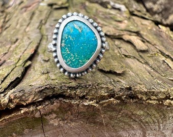 May include: A silver ring with a large, irregular turquoise stone. The stone is a vibrant mix of blue and green, set in a textured silver bezel. The ring has a beaded border and a simple band.