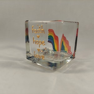 Op de afbeelding: Een glazen kaarsenhouder met een regenboogontwerp en de woorden "geloof, hoop, liefde" in goud aan de zijkant.