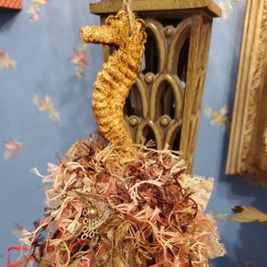 May include: A handcrafted seahorse decoration with a textured, golden-brown body. The seahorse hangs from a natural twine string, above a cluster of mixed fibres in shades of brown, gold, and coral. A decorative, vintage-style embellishment is attached to the fibre cluster.