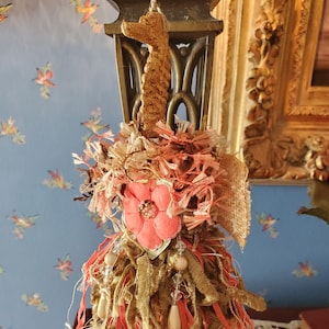 Könnte beinhalten: Handgefertigter Zieranhänger mit einem seepferdchenförmigen Aufsatz. Der Anhänger besteht aus rosa, goldenen und beigen Stoffen, verziert mit einer rosa Blume und hängenden Perlen. Der Hintergrund zeigt eine blaue Wand mit floralen Mustern.