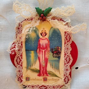 Imagen de tarjeta navideña vintage con ángel Art Deco decoupage sobre placa de madera pintada de rojo decorada con encaje y adorno de acebo, adorno único