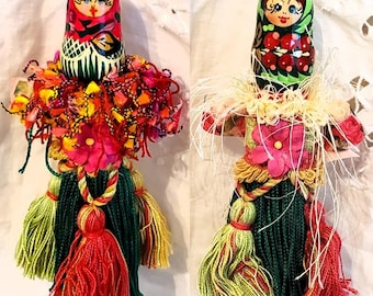 May include: Two Matryoshka doll ornaments. The dolls have painted faces and are adorned with colorful fabric and yarn. The dolls have green, red, yellow, and pink accents. Each doll has a tassel at the bottom.