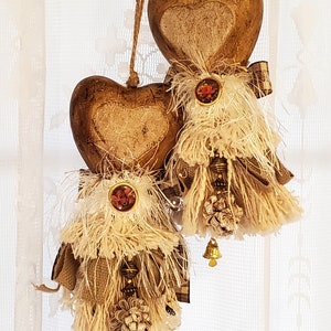 May include: Two hanging wooden heart ornaments with a distressed finish. Each heart has a smaller heart cutout and is adorned with fabric, fringe, and a small bell. The ornaments feature a rustic, handcrafted aesthetic, perfect for home decor.