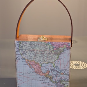 May include: A rectangular wooden handbag with a map design on the sides. The top is a darker wood, and it has a brown leather handle. The bag is secured with a gold-colored clasp.