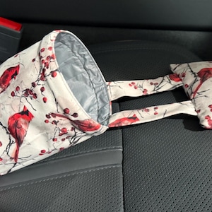 May include: A car trash bag and a small matching pillow, both featuring a white fabric with a red cardinal and berry design. The bag has a silver interior and is connected to the pillow by fabric straps. The items are placed on a black car seat.