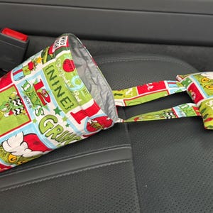 May include: A fabric car trash bag and a small matching pouch, both featuring a colorful Dr. Seuss Grinch-themed print. The bag is cone-shaped and open at the top, with a silver lining. The pouch has a strap. The items are on a black car seat.