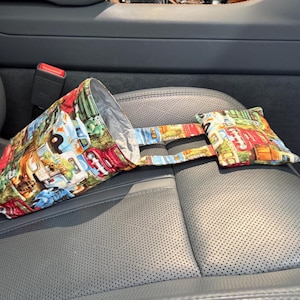 May include: A colorful fabric car trash can with a vintage truck pattern. The trash can is hanging on the back of a car seat.