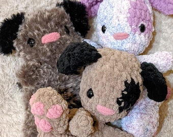 Puede incluir: Tres animales de peluche tejidos a crochet, un perro marrón, un perro blanco y morado y un perro marrón y blanco, están tumbados sobre una manta blanca esponjosa.