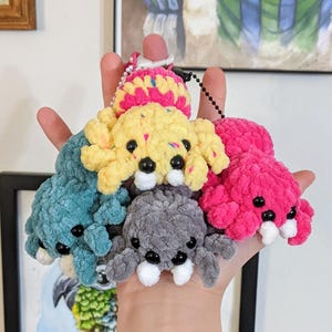 May include: A collection of colorful, crocheted spider-shaped keychains. The spiders are in shades of blue, yellow, pink, gray, and a multi-colored top. Each spider has black eyes and white fangs. The keychains are held in a hand.