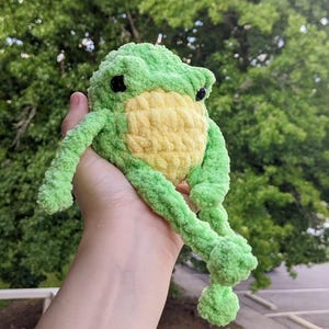 May include: A handmade, plush frog toy in shades of green and yellow. The frog has black eyes and is made of soft, textured yarn. The frog's body is a light green, with a yellow belly. The frog's arms and legs are long and floppy.