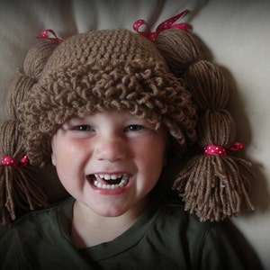 May include: A child wearing a brown crocheted hat with yarn pigtails tied with pink polka dot ribbons. The child is smiling and has a happy expression.