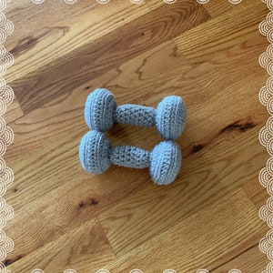 May include: A pair of gray crocheted dumbbells on a wooden surface.