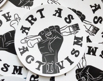 May include: A close-up of several round stickers with a black and white design. The stickers feature a raised fist holding paintbrushes and the text "ART IS ACTIVISM".
