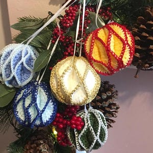 Op de afbeelding: Vijf gehaakte kerstballen in verschillende kleuren, waaronder blauw, wit, geel, rood en groen. De ballen hangen aan een kerstboomtak.