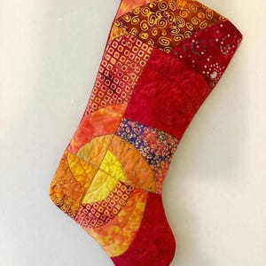 May include: A quilted Christmas stocking featuring a patchwork design of various fabrics in shades of red, orange, and yellow. The surface displays intricate patterns including swirls, dots, and geometric shapes, finished with a red fabric hanging loop.
