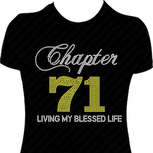 Puede incluir: Camiseta negra con letras de pedrería blanca que dicen "Chapter 71 Living My Blessed Life". Los números 71 están hechos de pedrería amarilla.