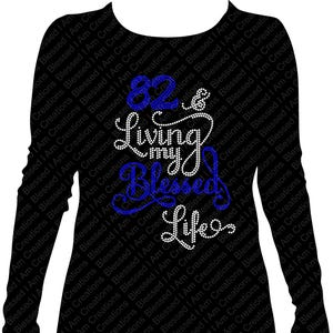 May include: Black long-sleeved shirt with the text "82 & Living my Blessed Life" in white and blue script. The shirt has a crew neck and a fitted silhouette. The design is centered on the front of the shirt.
