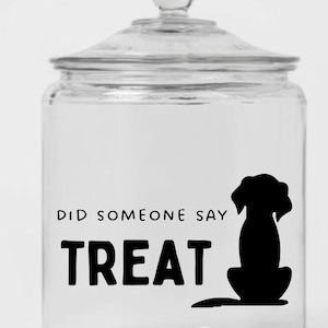 May include: A clear glass jar with a lid, featuring the text "DID SOMEONE SAY TREAT" in bold black letters. A black silhouette of a dog is on the right side of the jar. The jar is designed for storing dog treats.