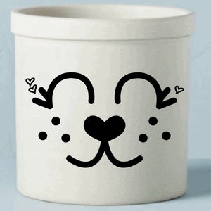 May include: A white ceramic container with a black cartoon dog face design. The design includes curved lines for the eyes, a heart-shaped nose, and dots for freckles. Small hearts are near the eyes.